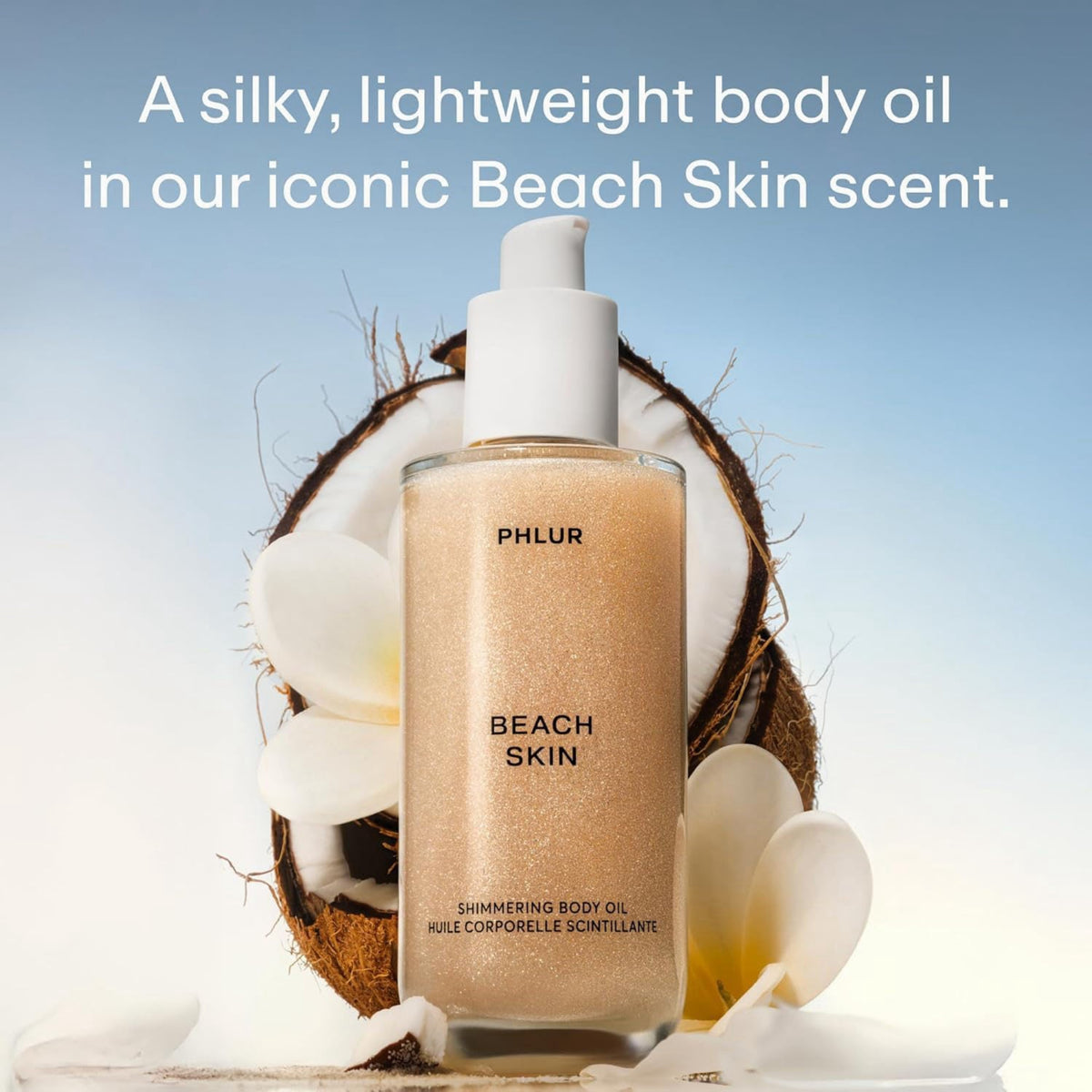 A bottle of PHLR Beach Skin shimmering body oil stands in front of a halved coconut and white flowers, with text at the top reading, 'A silky, lightweight body oil in our iconic Beach Skin scent.'.